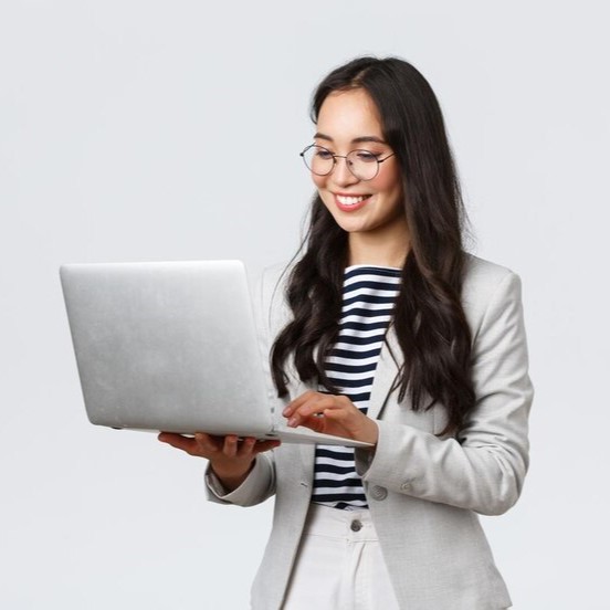 A happy Asian woman wearing a white blazer and glasses smiles as she looks at a silver laptop she is holding.